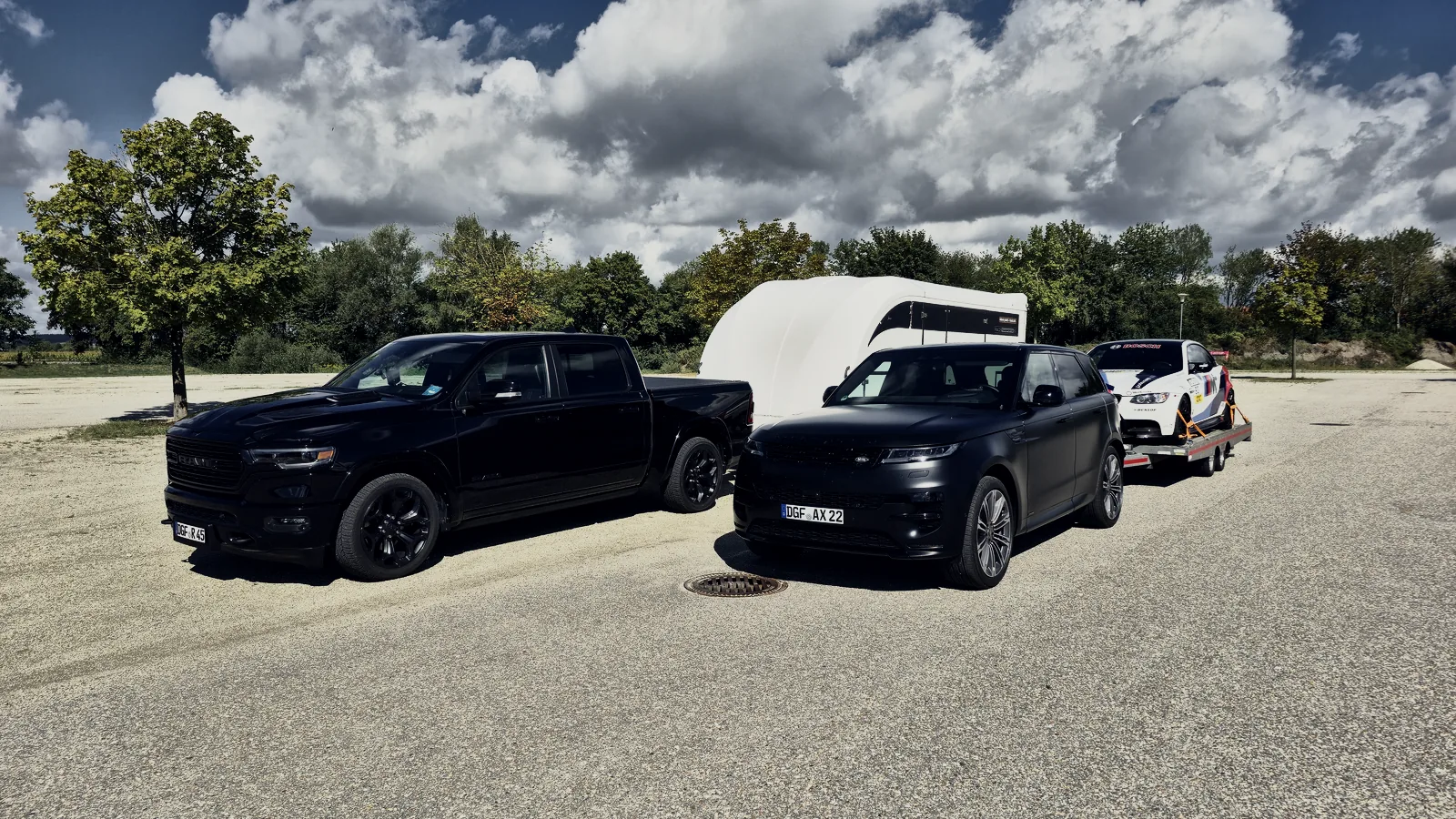 Zwei schwarze Zugfahrzeuge von AXX nebeneinander, dahinter der geschlossene Anhänger und ein BMW M3 auf der offenen Plattform